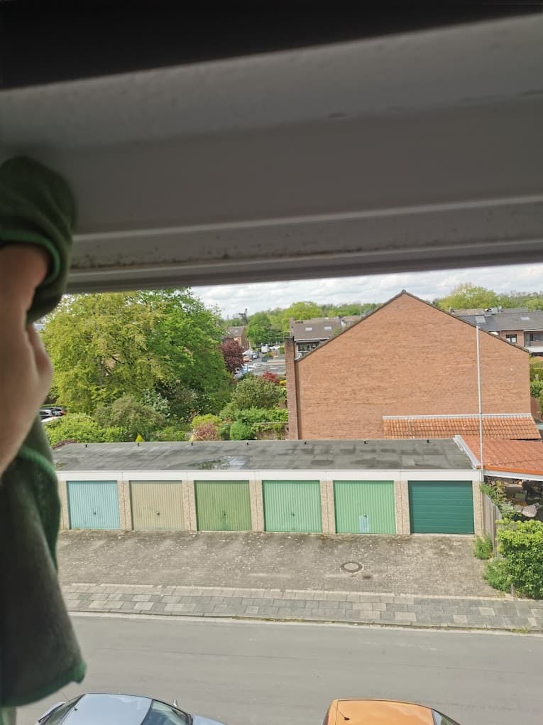 Ein Blick aus dem Fenster zeigt eine Reihe Garagen mit grünen Toren und ein Wohngebiet im Hintergrund. Eine Hand mit einem Putzlappen ist im Vordergrund zu sehen.