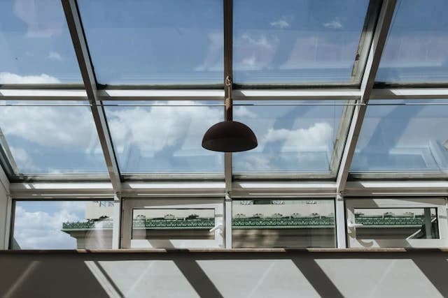 Ein Glasdach mit einer Lampe in der Mitte, durch das der blaue Himmel mit Wolken zu sehen ist. Das Dachfenster reflektiert ein Gebäude.